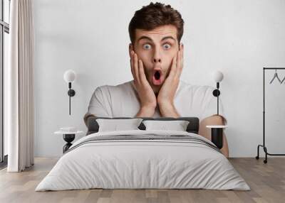 Portrait of shocked stupefied young male model with trendy hairdo looks with frightened expression, doesn`t expect to recieve news, keeps hands on cheeks, isolated over white concrete background Wall mural